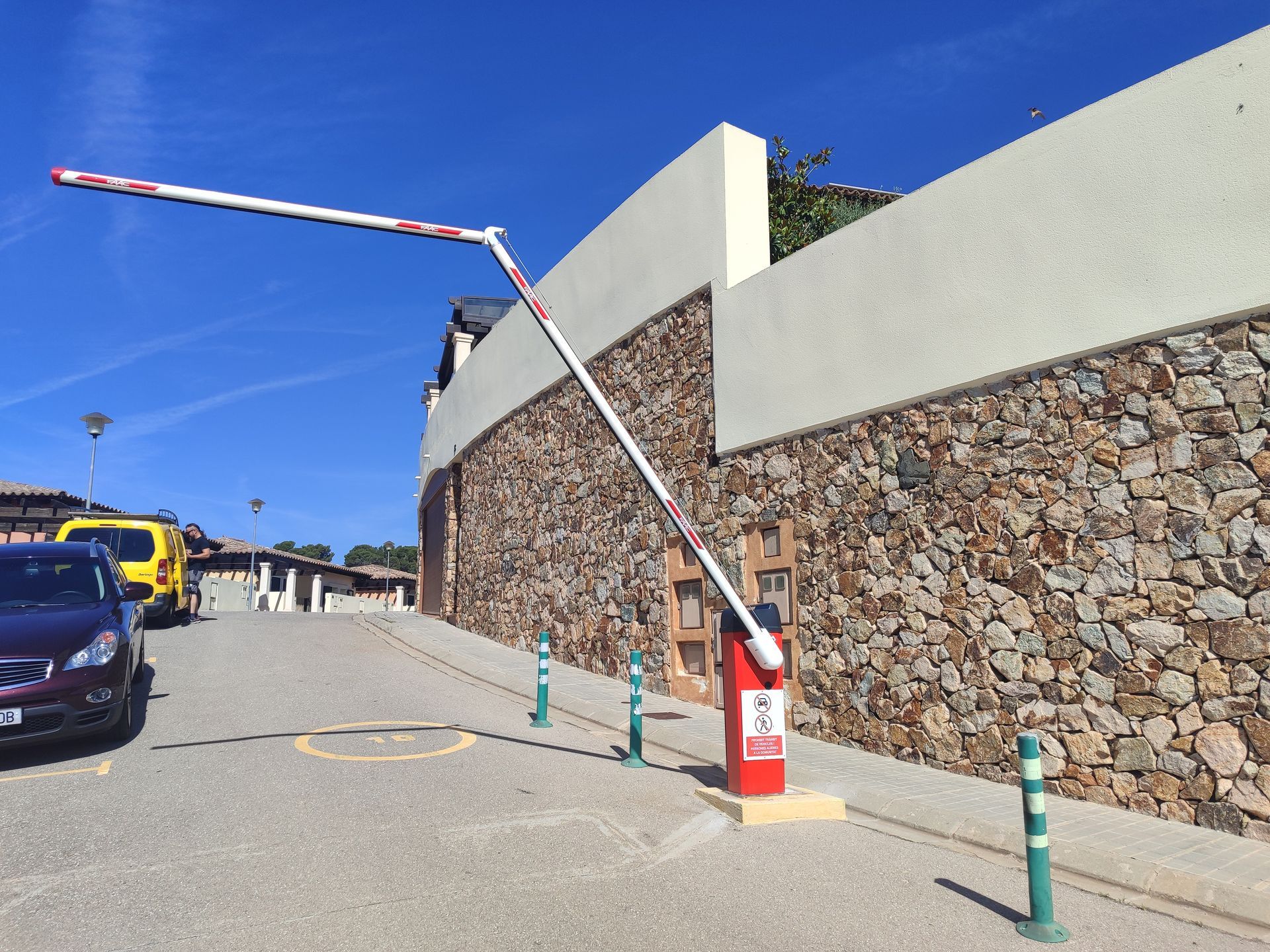 Una barrera a la entrada de un aparcamiento, sobre un muro de piedra. Un brazo rojo y blanco se levanta para permitir el acceso de vehículos.