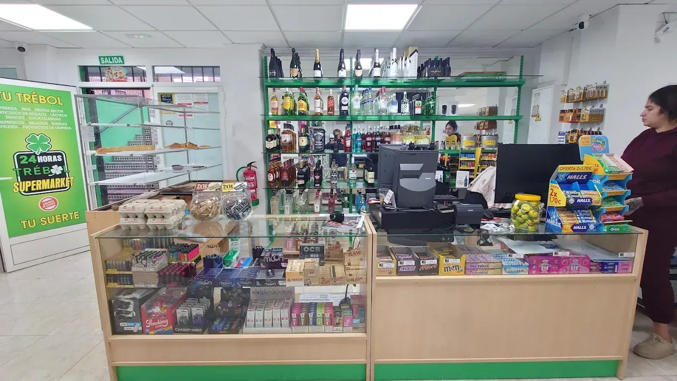 Dentro de una tienda de conveniencia, estantes repletos de productos, un empleado en la caja registradora.