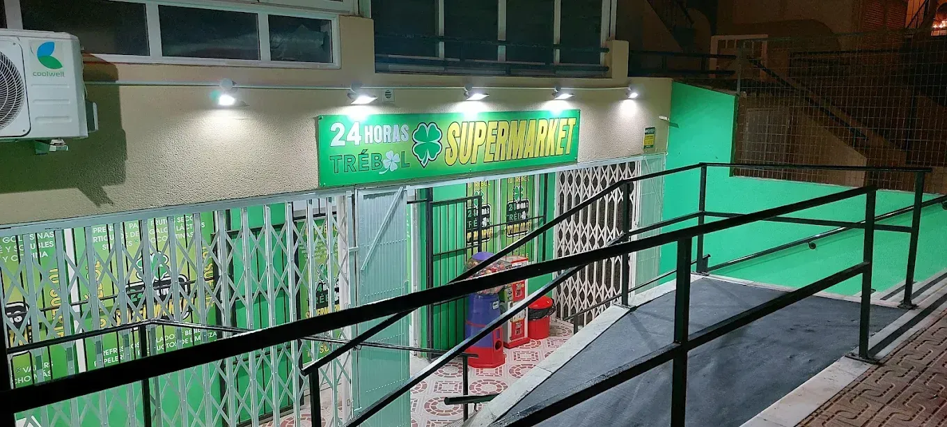 Vista exterior de una tienda de conveniencia abierta las 24 horas con un escaparate verde y blanco, una rampa y barandillas.