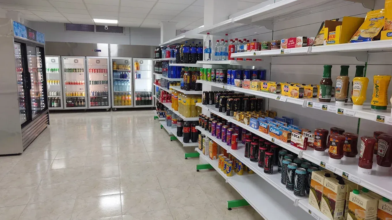 Un pasillo de una tienda de comestibles con estantes repletos de alimentos y bebidas; al fondo, vitrinas refrigeradas.