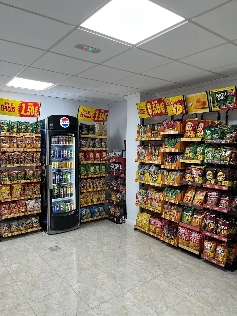 Dentro de una pequeña tienda de conveniencia, estantes llenos de patatas fritas y bebidas en una hielera.