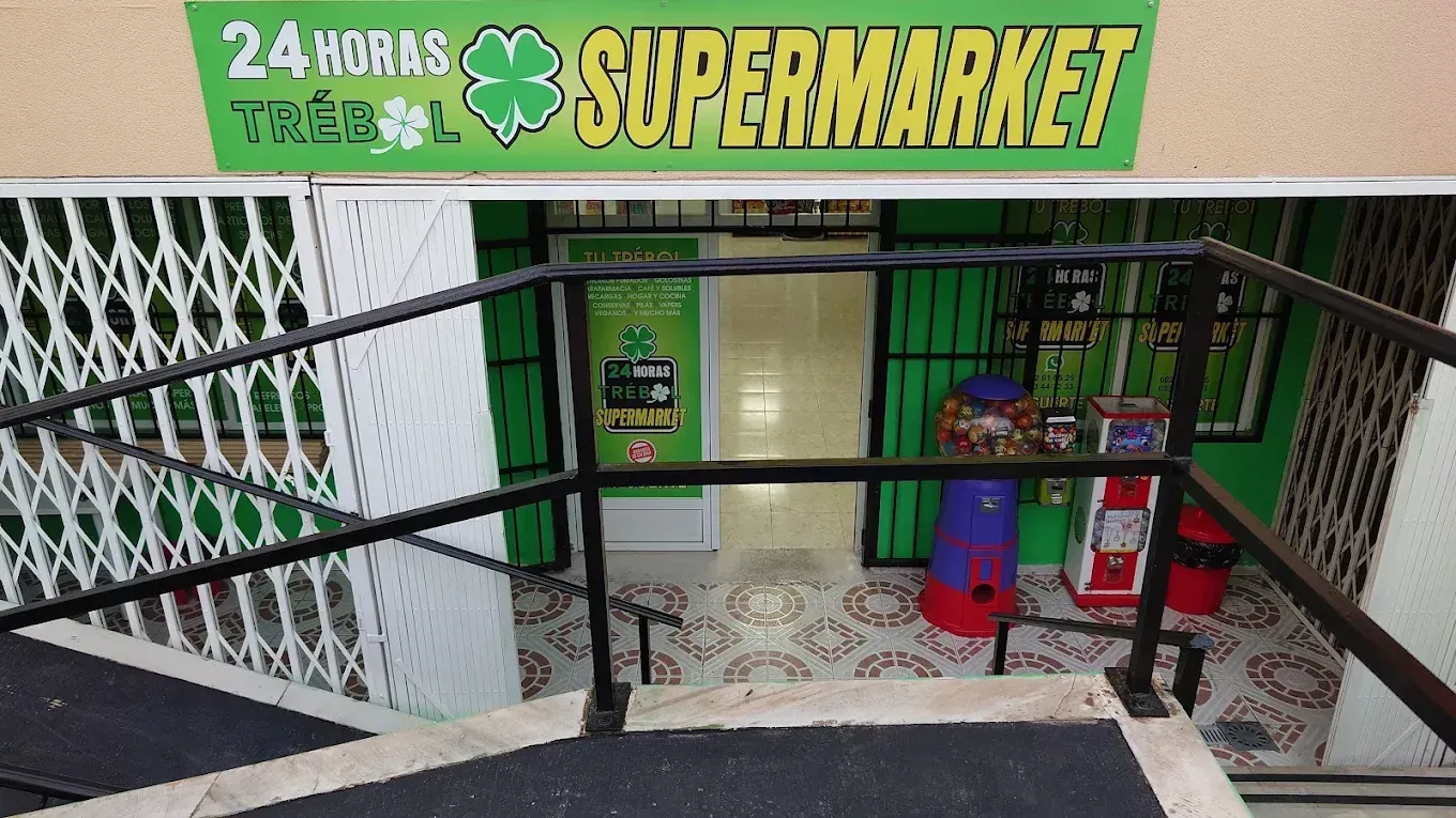 Entrada a un supermercado con un cartel verde y escalones. Se ven una puerta metálica y barandillas.