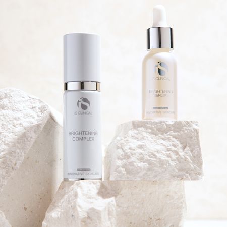 Zwei iS CLINICAL Hautpflegeflaschen, ein Brightening Complex und ein Brightening Serum, stehen auf strukturierten weißen Steinblöcken.