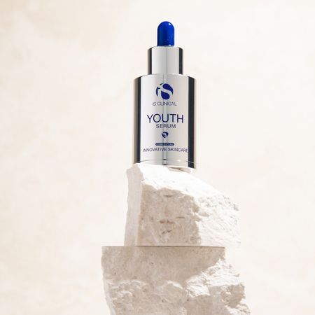 Eine silberne Flasche iS Clinical Youth Serum steht auf zwei übereinandergestapelten, rauen weißen Steinblöcken vor einem neutralen Hintergrund.