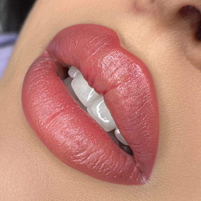 Nahaufnahme der Lippen mit einem weichen, gedeckten, matten rosa Lippenstift und sichtbarer Hautstruktur, hinter deren leicht geöffneten Lippen weiße Zähne zu sehen sind.