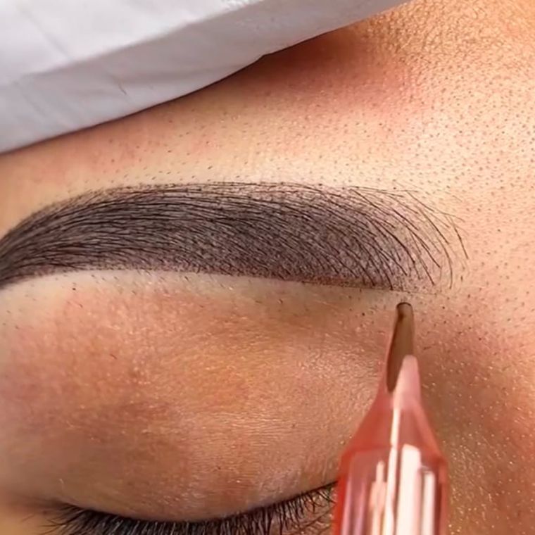 Nahaufnahme der Nadelspitze einer Tätowiermaschine, die Pigment aufträgt, um haarähnliche Striche auf einer Augenbraue zu erzeugen.