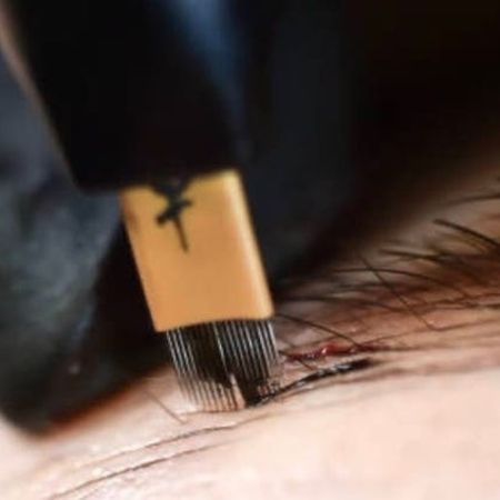 Nahaufnahme eines Microblading-Werkzeugs mit mehreren Nadeln, das dunkles Pigment in die menschliche Haut tätowiert.