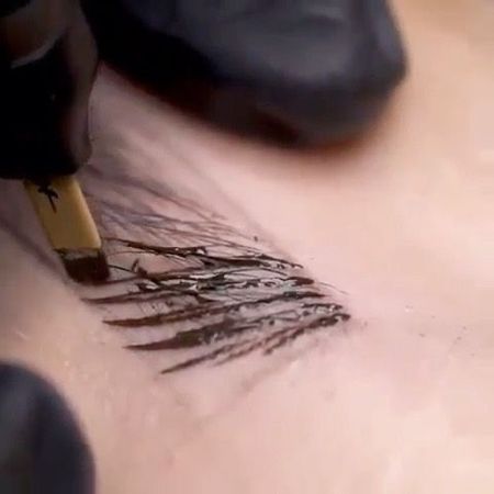 Mit einem behandschuhten Finger werden mithilfe eines Microblading-Werkzeugs haarähnliche Striche auf der Haut erzeugt, um die Augenbrauen zu formen.