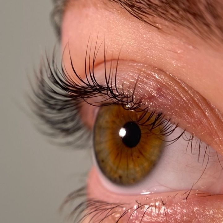 Eine seitliche Nahaufnahme eines menschlichen Auges mit haselnussbrauner Farbe und langen, geschwungenen dunklen Wimpern.