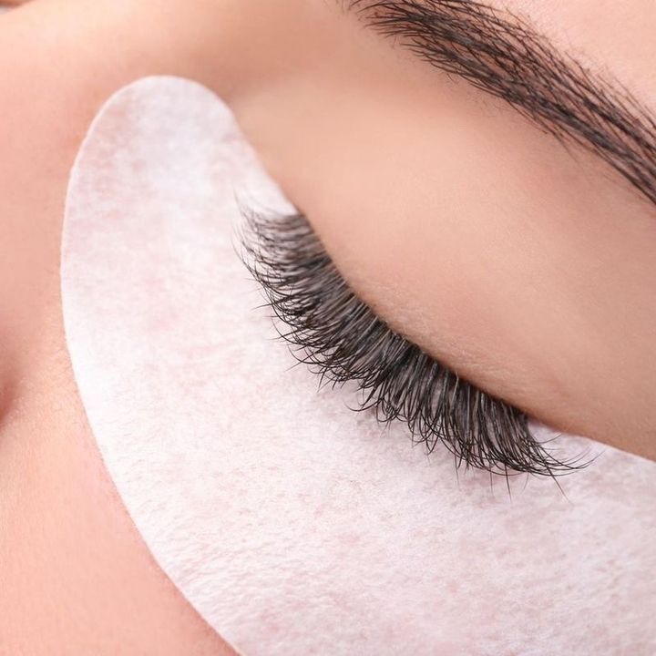 Nahaufnahme eines geschlossenen Auges mit dunklen Wimpernverlängerungen und einem weißen Augenpad, das auf die Haut unter dem Auge geklebt wurde.