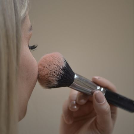 Eine Person hält einen großen, flauschigen Make-up-Pinsel mit rosafarbenen Borstenspitzen an ihre Wange.