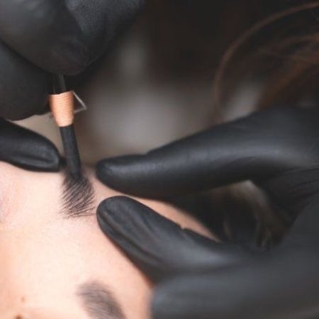 Eine Technikerin in schwarzen Handschuhen trägt mit einem Microblading-Werkzeug manuell Pigmente in die Augenbrauen der Kundin ein.