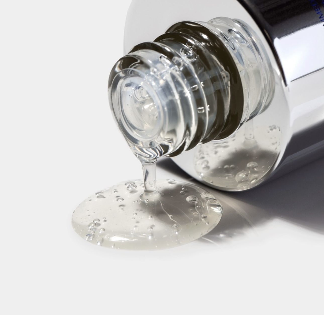 Eine klare, zähflüssige Flüssigkeit tropft aus der Öffnung einer silbermetallischen Hautpflegeflasche auf eine weiße Oberfläche.