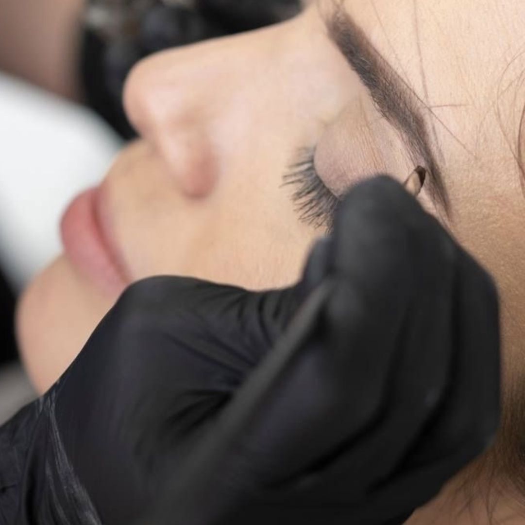 Eine Person in schwarzen Handschuhen trägt mit einem Kosmetikinstrument präzise Make-up oder Microblading im Bereich der Augenlider einer Kundin auf.