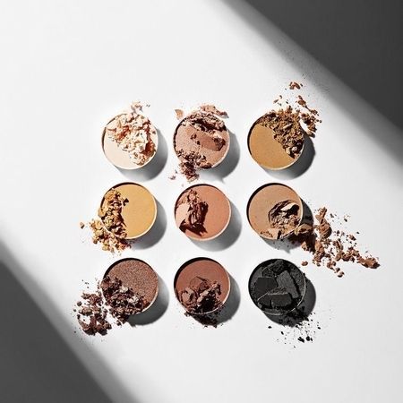 Neun runde Lidschattenpfännchen, die in einem Raster angeordnet sind und von Beige über Dunkelbraun bis Schwarz reichen, mit sichtbarem losem Puder.