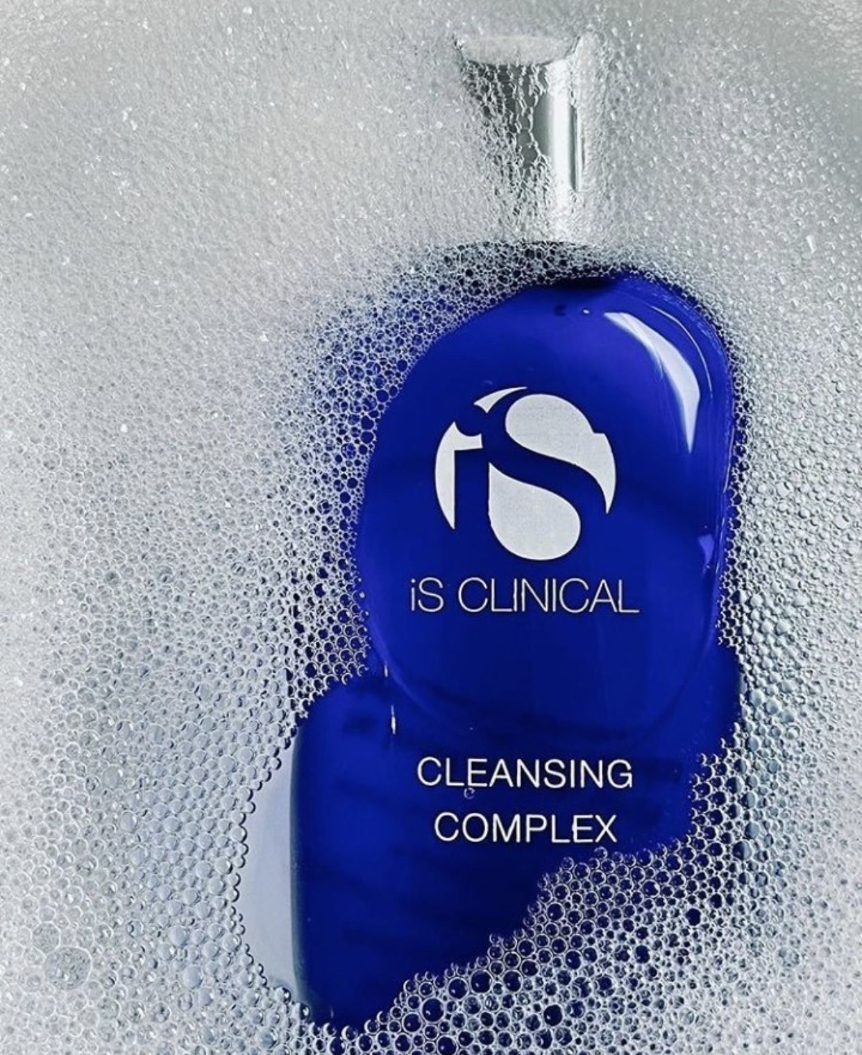 Eine Flasche iS CLINICAL Cleansing Complex, eingebettet in reichhaltige, weiße Seifenblasen.