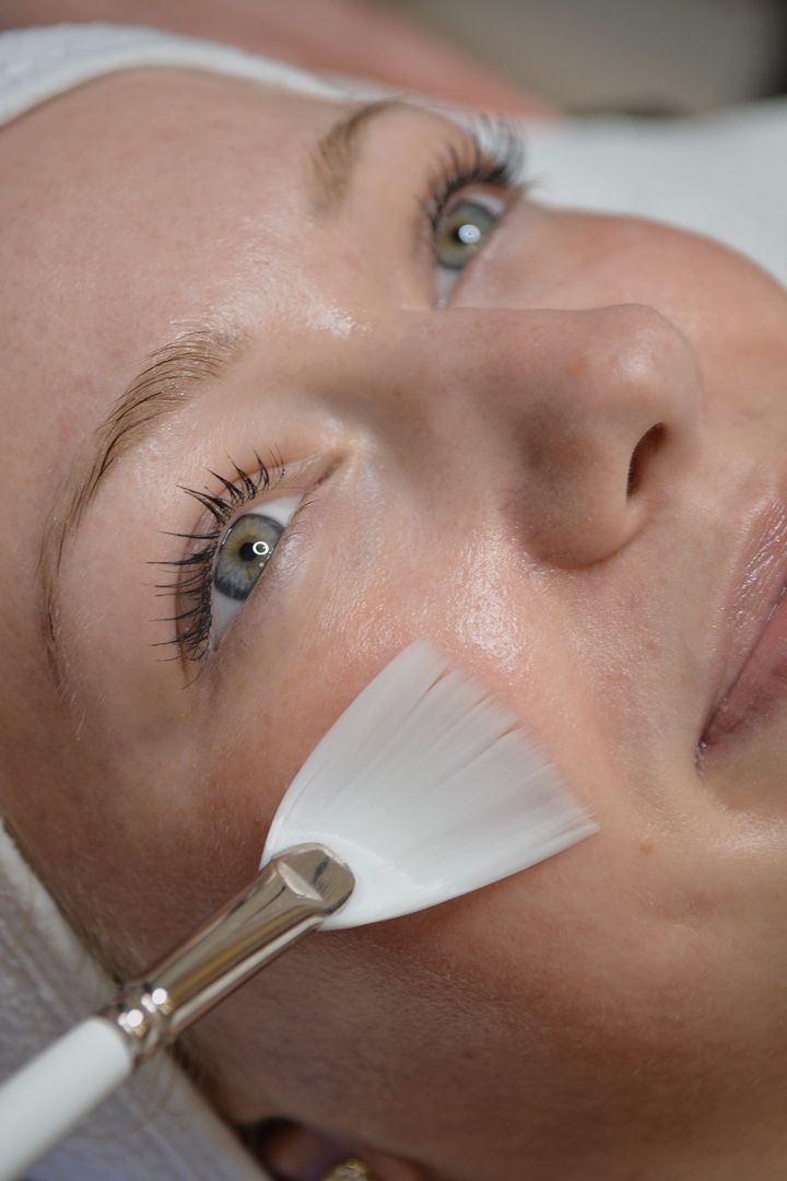 Eine Person liegt auf einer Behandlungsliege und erhält eine Gesichtsbehandlung, bei der mit einem weißen Fächerpinsel ein Produkt auf die Wange aufgetragen wird.
