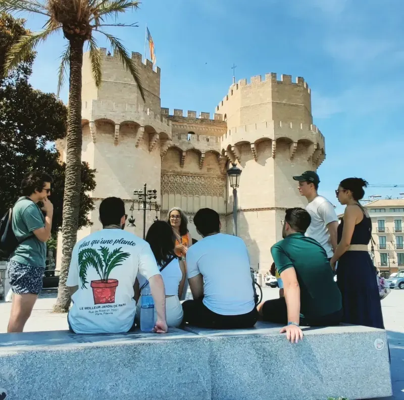Gente sentada en un banco frente a un castillo de color crema bajo un cielo azul, con palmeras cerca.