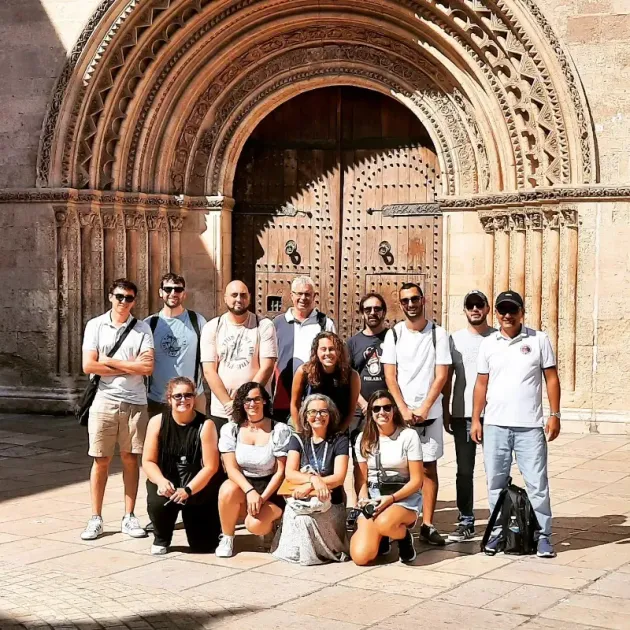Grupo de turistas posando frente a una puerta de madera arqueada y ornamentada en un soleado patio de piedra.