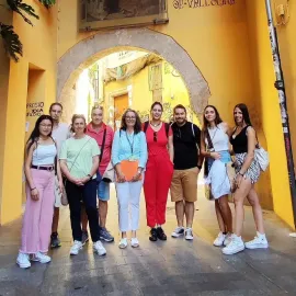 Un grupo de ocho personas posando bajo un arco en una calle estrecha, soleada y de color amarillo.