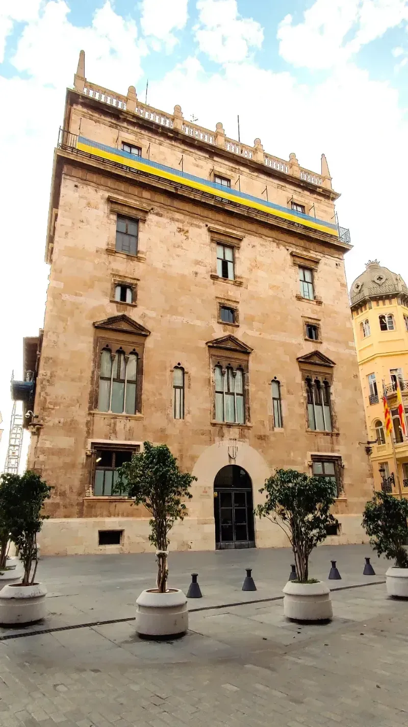 Edificio histórico de piedra con ventanas arqueadas y un balcón amarillo en una plaza soleada de la ciudad.
