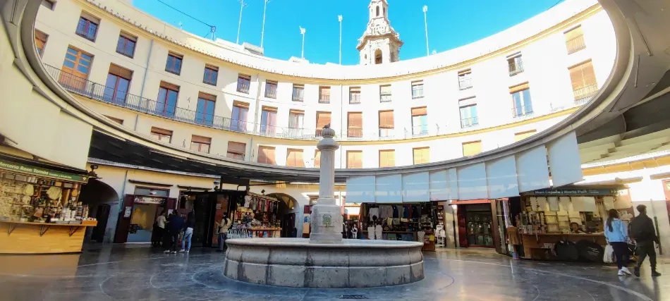 Vista panorámica de una plaza soleada con una fuente central bajo la fachada curva de un edificio y un reloj de torre.