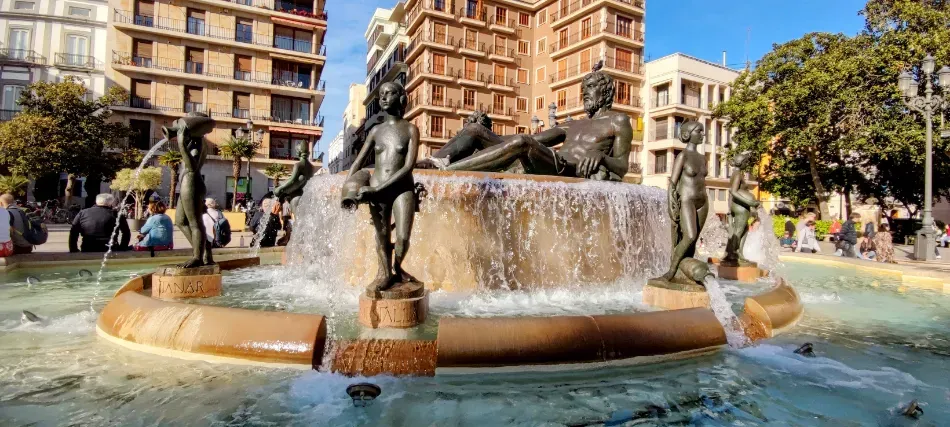 Fuente con estatuas de bronce en una plaza de la ciudad, agua que cae en cascada a una piscina, rodeada de edificios y árboles.
