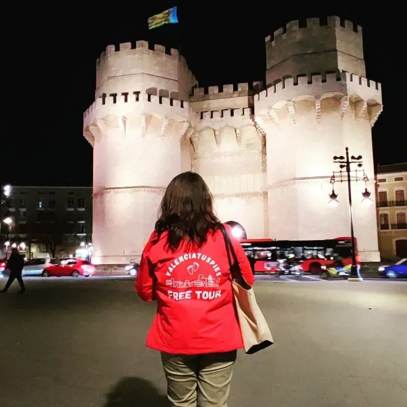 Una persona con una camisa roja se encuentra de pie frente a una histórica puerta de piedra iluminada por la noche.