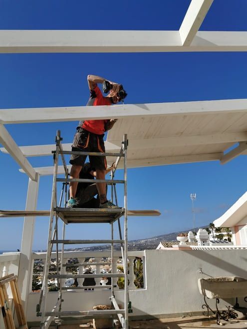 Un trabajador en un andamio instala una viga de techo blanca bajo un cielo azul.