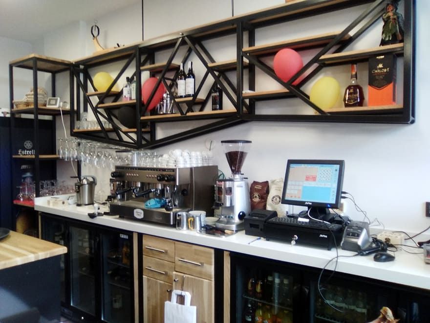 Cafetería moderna con máquinas de espresso, molinillo, estanterías y una estación de trabajo informática detrás del mostrador.