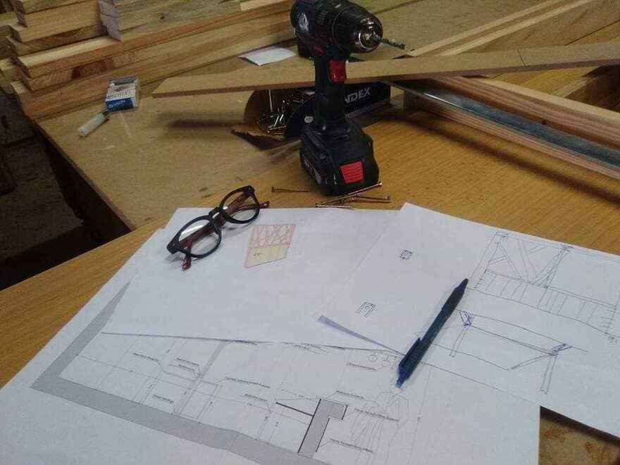 Taladro inalámbrico sobre un banco de trabajo junto a unas gafas y un lápiz sobre planos de construcción y tablas de madera.
