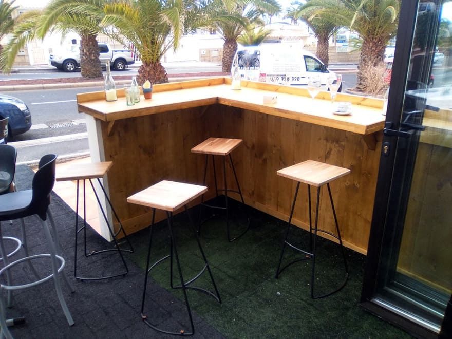 Terraza de una cafetería con barra de madera, taburetes altos y palmeras al fondo.
