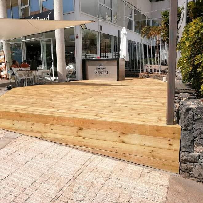Plataforma elevada de madera frente a un restaurante junto al mar con terraza y un muro de piedra.