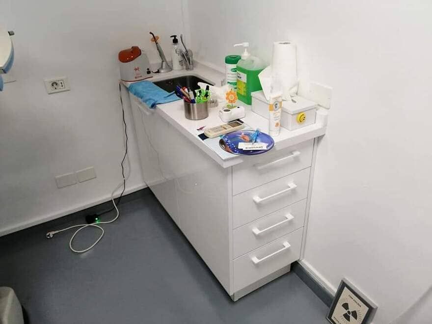 Puesto de tratamiento de clínica dental con mueble blanco, lavabo, instrumental y suministros sobre un suelo gris.