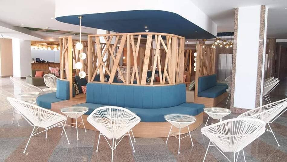 Salón moderno con asientos azules, sillas de alambre blancas y una mampara circular de madera bajo un techo curvo.