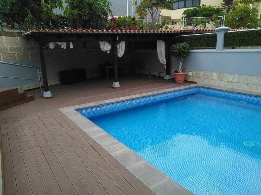 Piscina en el patio trasero con agua azul, terraza de baldosas y zona de estar con sombra.