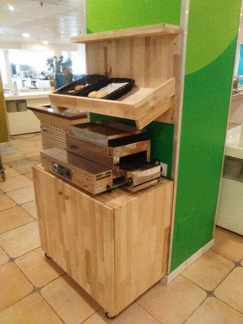 Expositor de madera para alimentos con bandejas y cajas de pasteles en una luminosa cafetería o mercado interior.