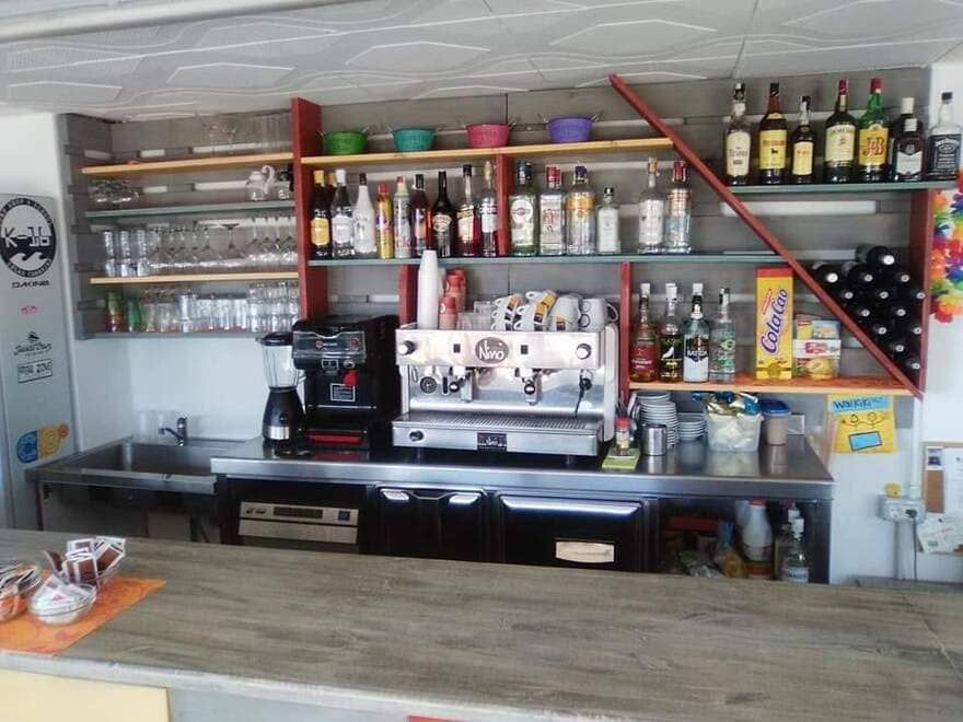 Barra con máquina de espresso, cristalería, botellas y estantes con bebidas en un ambiente de cafetería.