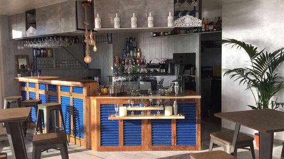 Interior acogedor de bar con barra de madera, paneles azules, taburetes, lámparas colgantes y plantas.