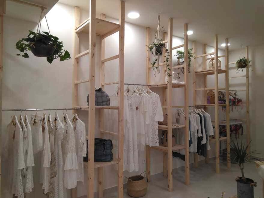Boutique de ropa minimalista con prendas blancas en percheros de madera y plantas colgantes.