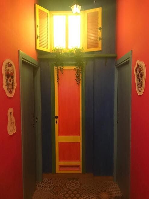 Pasillo colorido con paredes azules y coral, suelo estampado, ventana luminosa y vegetación colgante sobre una puerta roja.