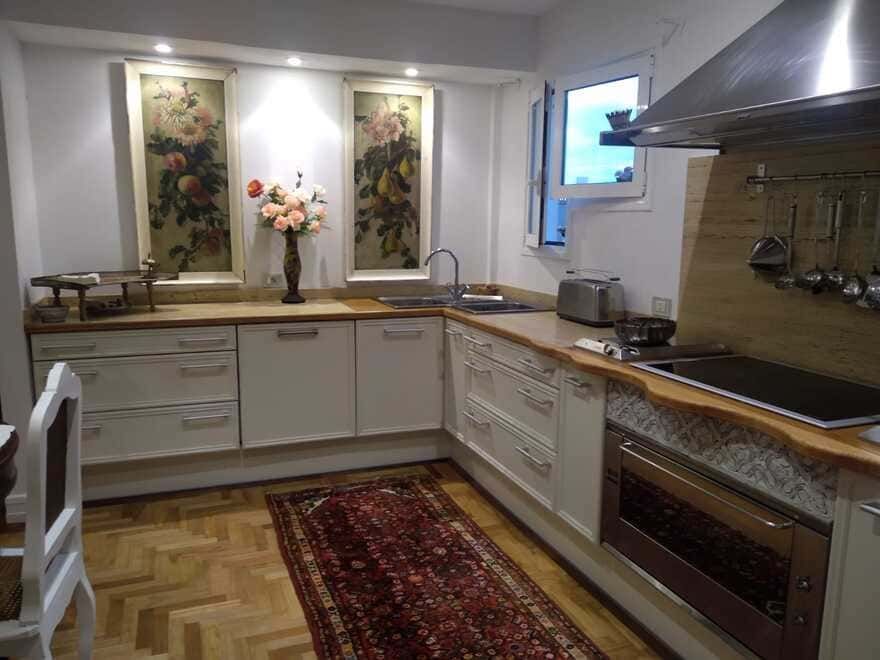 Cocina compacta con armarios blancos, encimeras de madera, decoración floral en la pared y una alfombra roja alargada.