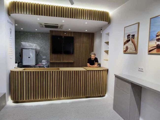 Mostrador de recepción moderno con listones de madera, televisor y un miembro del personal de pie detrás en un vestíbulo luminoso.