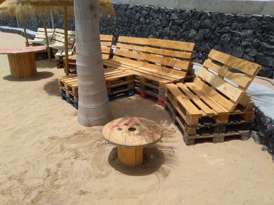 Banco de madera curvado alrededor de una palmera en una playa de arena junto a un muro de piedra y una mesa pequeña.