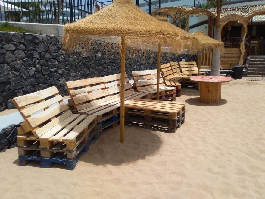 Zona de descanso en la playa con bancos de madera, sombrillas de paja y suelo de arena junto a un muro de piedra.