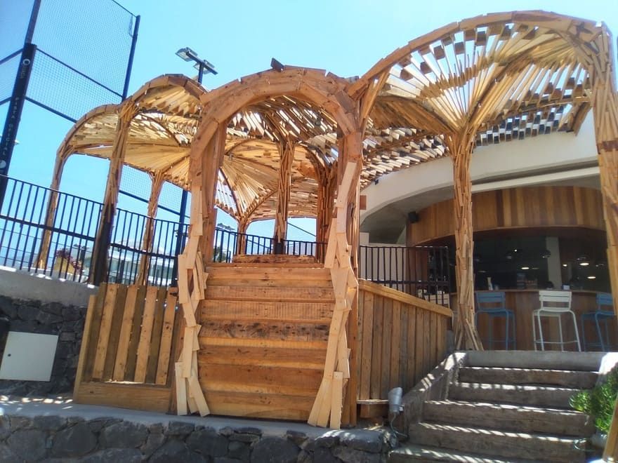Escalera de madera y entrada con pérgola y arcos tallados junto a un edificio bajo un cielo azul.
