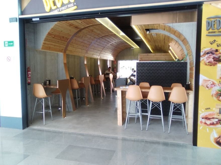 Zona de estar vacía de la cafetería con sillas beige bajo un techo de madera curvado, vista desde la entrada.
