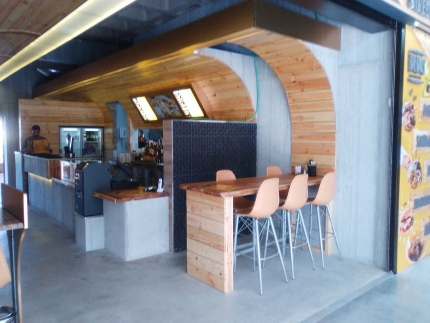 Interior de cafetería moderno con barra de madera y metal, taburetes altos y máquinas de espresso.