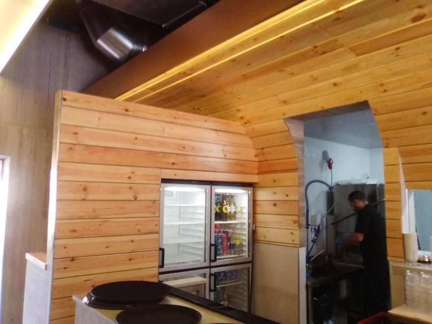 Zona de cocina con paneles de madera, un frigorífico con puerta de cristal y una persona trabajando cerca de una estufa.