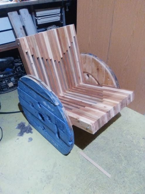 Silla de madera con asiento y respaldo de listones, soporte lateral azul, en un entorno de taller.